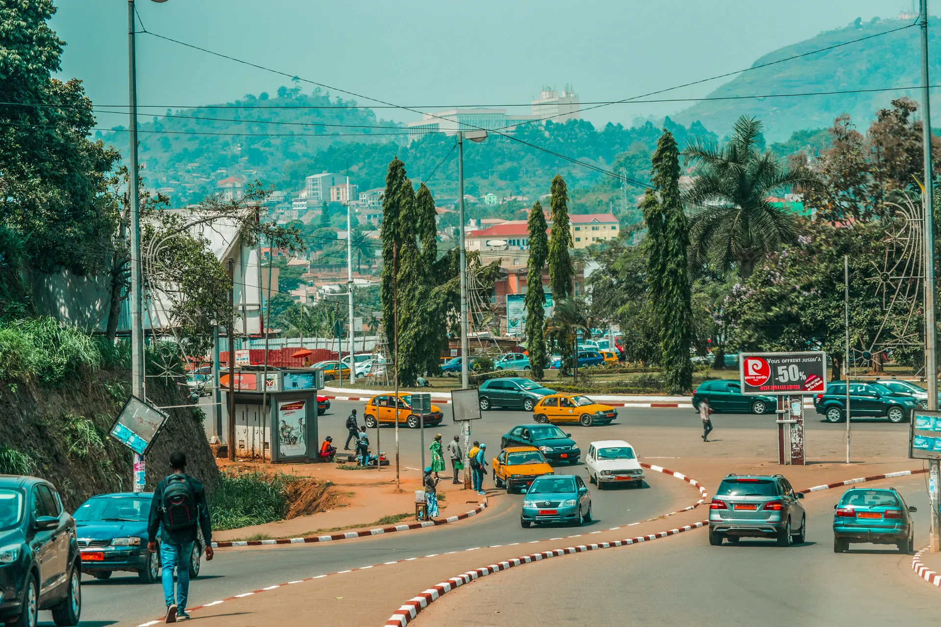 Street scene in Yaoundé, Cameroon, showing a busy curved roadway with cars and yellow taxis circling a roundabout.