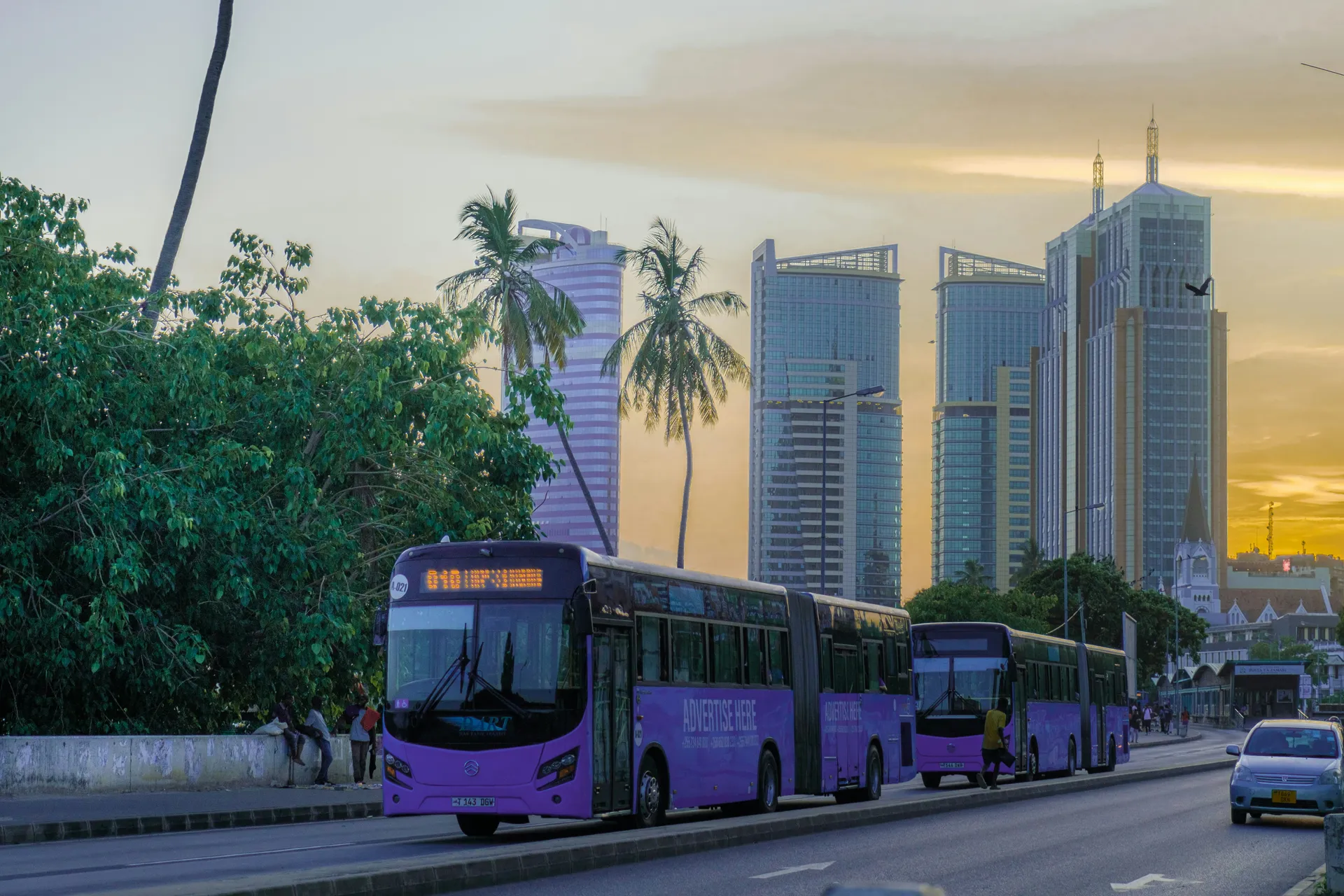 Purple rapid transit buses travel along a road in Dar es Salaam, Tanzania, with palm trees and modern glass skyscrapers against a golden sunset sky.